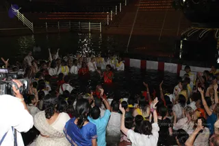 Night photo of a large crowd gathered in and around a stepped water tank lit by strings of lights. In the water, a group of devotees in white and yellow-trimmed white garments stand waist-deep, and a woman in an orange-red sari stands centre, arms raised. Behind her, a figure in red-orange robes with a red-and-orange garland stands in the water. Many people on the steps and in the water raise their hands in devotion. At left, a cameraman films the scene. Garlands of flowers decorate the edge at right. The crowd is dense and visible across the frame, so the count is well above twenty.