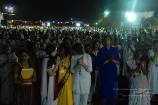 Night outdoor gathering of a very large crowd facing a distant stage, lit by floodlights and strings of colourful bulbs. In the foreground, several women stand with hands pressed together in namaste or prayer. A woman in a bright yellow sari with a gold border holds a small badge. A woman in white salwar with a white dupatta and a rudraksha-style mala presses her palms together. A woman in a deep blue shawl stands with hands folded in front of her chest. Behind them, many more people in white and light clothing stand in rows along wooden barriers, some wearing yellow and red lanyards. A large purple bird-shaped emblem stands on a tower at left. The scene suggests a satsang or public discourse, with the crowd standing in devotional attention.