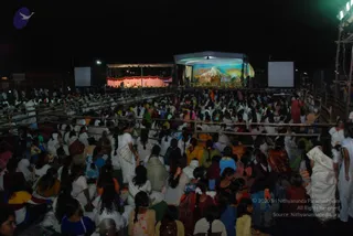 Night-time outdoor gathering of a large crowd, mostly women, seated on the ground behind bamboo barricades facing a stage. Many wear white saris and salwar kameez in colours such as yellow, orange, pink, green and blue, with some hair adorned with flowers. On the stage, a backdrop painting shows a snowy mountain peak under a pale sky, and figures sit in the lit area below. Large white projection screens flank the stage, and bright floodlights stand at the right edge. A few standing figures in white move among the seated crowd. Crowd size is too large to count precisely.