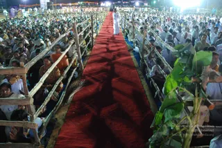 Night photograph from an elevated angle of a large outdoor gathering in front of a stage. A long red carpet runs down the centre between rows of bamboo-railed barriers. At the far end of the carpet, a lone figure in white clothing stands. Seated devotees on both sides, many in white and some women in saffron and orange saris, face the carpet. Green banana leaves appear at the right edge, and string lights and bright stage lights glow in the background. Visible people are numerous; the crowd is large, so the count range is 100+. No clear identification of the figure at the end of the carpet is made from appearance; he is described only by role as a person in white standing at the stage end.