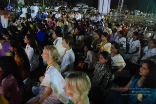 Night photo of a large open-air gathering of devotees seated on the ground in rows, facing toward the right. Many wear white or cream clothing with gold-bordered saris and mustard or yellow lanyards with badges. Women in blue, purple, and patterned dark saris sit in the foreground, one woman with a blonde ponytail in a white sari with a gold border. Men in white shirts sit among them in the background. A wooden staging and white banner sit at the upper right, with lights and vehicles beyond. No a person candidate is visible.