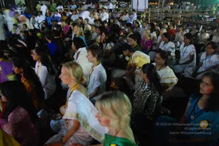 A night-time outdoor gathering of many devotees seated on the ground in rows, facing toward the front. Most wear white or light traditional clothing, including cream and gold-bordered saris and white kurtas, with lanyards and identity badges around their necks; a few wear bright blue, pink, green and dark embroidered outfits. Some have eyes closed in meditation. Bamboo scaffolding and tents frame the background, with lamps glowing at the top right. No a person figure is visible in the frame, so the scene shows an audience rather than a presenter.