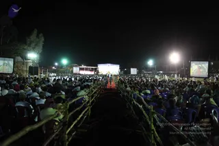 A night-time outdoor event photographed from the back of a seated audience. Hundreds of people sit in rows of blue plastic chairs on both sides of a central aisle lined with bamboo railings, with a red carpet running toward a lit stage at the far end. Several large screens flank the audience and the stage, displaying images, and floodlights and green lamps illuminate the field. Distant figures in light clothing walk near the stage. Most people visible are seated at a distance, many wearing white or light garments, with some heads covered by cloths.