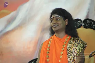 A photograph of a man seated in a dark carved wooden chair against a large painted backdrop of pale mountain and orange sky. He has long dark hair, a white forehead marking with a red dot, and wears an orange saffron robe with a leopard-print shawl over one shoulder and a long gold bead necklace. Swamiji (probable): the long swept-back hair, saffron robe, ornate gold beaded necklace and shawl are consistent with the page's reference to a talk, and he is seated as the speaker.