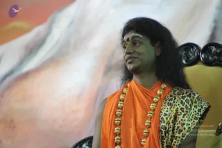 A man with long dark hair and a red bindi on his forehead sits on an ornately carved dark wooden chair, turning his head to the side. He wears an orange saffron wrap draped over his shoulders, a leopard-print shawl over one arm, and a long multi-strand necklace of gold beads hanging to his chest. A large painted backdrop of soft orange, pink and white clouds or mountains fills the background. A microphone is visible at the lower right edge. Swamiji (probable): long swept-back hair, saffron clothing and ceremonial gold beaded necklace, seated in a chair at a satsang stage.