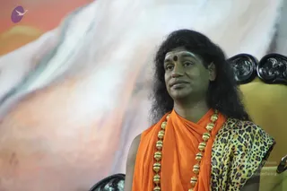 A man with long, swept-back dark hair and a red bindi sits on an ornate black carved chair before a large painted backdrop of pale peaks and orange sky. He wears an orange saffron shawl over a leopard-print garment and a long multi-strand necklace of large gold beads. Swamiji (probable): long swept-back hair, saffron clothing and ceremonial gold beads, consistent with the page's reference to a Shivaratri satsang discourse. He appears to be seated in a speaker's position, looking off to one side.