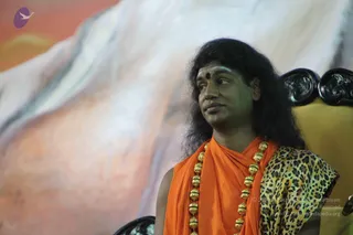 A man with long, swept-back dark hair and a red bindi stands before a large painted backdrop of pale mountains against orange sky. He wears an orange saffron shawl draped over the shoulders and a leopard-print garment, with a long ceremonial gold bead necklace. He looks off to the side toward a carved dark wooden chair. The appearance matches the probable Swamiji profile, with long hair, saffron clothing and gold bead garland cues, and he is the only person visible.
