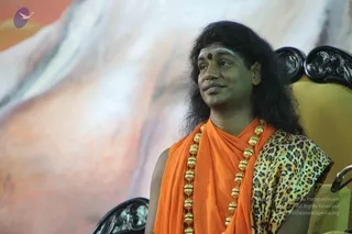 A man with long dark hair and a red bindi sits in a carved black-and-gold throne-style chair against a soft orange and white painted backdrop. He wears an orange saffron wrap over a leopard-print garment and a long necklace of large gold beads. He gazes to the side with a slight smile. Swamiji (probable): long swept-back hair, saffron wrap and ornate gold beaded necklace.