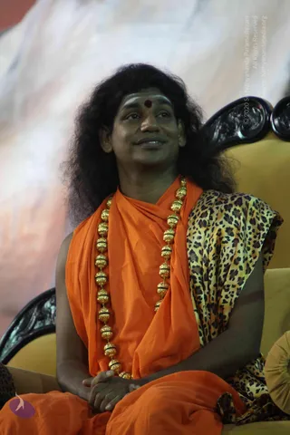 A seated man with long, dark, swept-back hair sits in a carved dark-wood chair with yellow upholstery. He wears an orange-saffron shawl over his shoulders, a leopard-print wrap draped over one arm, and a heavy ornate gold bead necklace of large round beads. He has a red bindi and light ash marks on his forehead, and his hands rest clasped in his lap as he gazes upward with a gentle smile. Appearance-based candidate: a person, shown in person in a seated stage or satsang setting with a pale backdrop.
