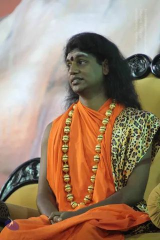 A seated man with long, swept-back dark hair wears an orange saffron wrap over a leopard-print shawl and a long ceremonial gold bead necklace, with a red bindi on his forehead. He sits in a yellow gilded chair with carved black armrest, hands folded in his lap, looking to the side while speaking. A pale, blurred backdrop fills the background. Swamiji (probable): the long swept-back hair, saffron clothing, gold beaded necklace and leopard shawl are consistent with the page's claim that he is present at the event.