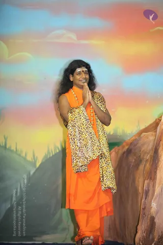 A long-haired man stands barefoot in front of a painted backdrop of a sunset sky with cacti and rocky hills. He wears an orange sleeveless robe with a leopard-print shawl draped over his shoulders and a gold bead necklace, a white tilak with a red dot on his forehead. He stands with palms pressed together in namaste and smiles. a person, matching long swept hair, saffron robe, leopard-print shawl and gold necklace cues, shown as a single person in a photo.