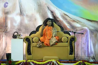 A photograph of a seated man on a stage giving a talk. He wears an orange saffron robe over a leopard-print garment and a long necklace of large gold beads, and has long dark hair and a red forehead mark. He sits on an ornate gold and black carved throne with round cushions, one hand raised in gesture, facing a microphone on a stand. A large painted backdrop of rocky mountain forms and sky stretches behind him, and the stage edge is decorated with marigold and orange flower garlands. A white pedestal with a small audio device sits to the left.