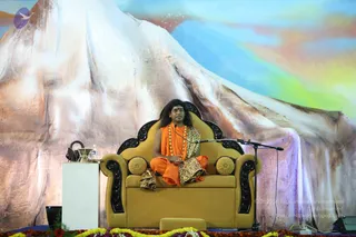 A photograph of a seated speaker on a golden-yellow upholstered armchair with carved black wooden trim and gold cushions, centered on a stage. He wears saffron-orange robes, a leopard-print shawl draped over his lap, and a multi-strand orange and white beaded mala around his neck, with long hair and a tilak on his forehead. He sits cross-legged and speaks into a boom microphone. Behind him is a large painted backdrop of a snow-capped mountain against a pastel sky. In front of the chair are marigold and yellow flower garlands along the stage edge, a white podium with a small glass vessel and microphone, and a black case. Swamiji (probable): long swept-back hair, saffron robes and a rudraksha-style bead mala identify this figure as a.