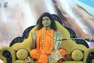 A seated man speaks into a microphone on a gold-upholstered, black-carved throne-like sofa against a painted mountain backdrop. He has long dark hair, a red tilak on his forehead, a saffron-orange robe, a heavy gold bead necklace, and a leopard-print shawl draped over his shoulders and lap. His right hand is raised in a gesture of explanation while his left rests on his knee. Swamiji (probable): long swept-back hair, saffron clothing, gold bead necklace and a ceremonial seated position on the throne, consistent with a discourse setting.