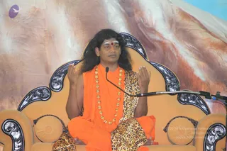 A man with long dark hair sits cross-legged on an ornate peach-coloured throne with dark carved armrests and round cushions. He wears an orange saffron robe draped over a leopard-print shawl and a long necklace of gold beads, with a red bindi and white vibhuti on his forehead. His hands are raised in front of him in a teaching gesture, and a microphone stand is positioned beside him. Behind him is a large painted backdrop of rocky mountain terrain in pink and cream tones. The scene appears to be a satsang discourse.