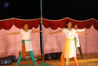 Three young women in a stage performance under a red and pink draped tent backdrop at night, standing in a wide-armed yoga pose on green and orange mats. The front woman wears a white kurta with a yellow and orange sash and orange trousers and a bindi; the woman on the left wears a white top with an orange and teal sash and teal trousers; the woman at the back wears white with a teal sash. Bags and luggage sit at the stage edge.
