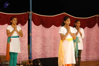 Three young girls stand in a row on a stage in front of draped pink and maroon fabric with scalloped valances. They are in prayer pose with palms pressed together at chest height. The girl on the left wears a white top, teal leggings and an orange and teal scarf. The centre girl wears a white top and long white skirt with a yellow and orange sash and a small red bindi. The girl on the right wears a white top with a teal and green shawl and has her hair tied back. A blue metal pole stands between them, and equipment cases sit at the stage edge.
