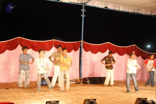 A night stage performance with a group of boys dancing on a sandy-floored stage under a canopy. Six boys are visible in casual clothes: a striped blue and white shirt with jeans, a white shirt with jeans and a red bead necklace, a khaki shirt with white trousers, a yellow shirt with white trousers, a dark patterned shirt with white trousers, and a white jacket with jeans. The backdrop is draped in red and pink fabric with scalloped valances, and dark stage lights are visible at the bottom edge. No a person candidate is visible.