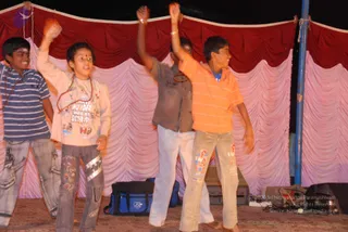 A night stage performance photograph showing four boys performing with raised arms in front of draped red and pink curtains. The boy on the far left wears a blue striped polo shirt and jeans; the next boy wears a light pink long-sleeved top printed with cartoon motifs, grey jeans and a red bindi and rudraksha-style beads; the third wears a grey-brown shirt and white trousers; the boy on the right wears an orange striped shirt and light patterned trousers. Blue and black equipment bags and speaker cases sit on the stage floor, and a blue pole stands at the right edge.