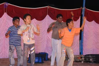 Four boys perform on a stage in front of a draped backdrop of red valance above pink gathered curtains, with a blue metal pole at the right edge. From left: a boy in a blue-and-white striped shirt and jeans, a boy in a cream jacket with a red-and-white printed design and a beaded necklace, a boy in a grey-brown shirt and white trousers with a light forehead mark, and a boy in an orange-and-white striped shirt with patched jeans, arms raised. Blue bags sit on the stage floor behind them.