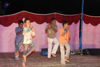 Four children perform on a stage at night in front of a draped backdrop of red and pink fabric. Each stands on one leg with palms pressed together in anjali mudra. The boy on the left wears a blue striped shirt and grey jeans, the girl next to him a cream top and patterned leggings, the boy in the center a brown shirt and white trousers, and the boy on the right an orange t-shirt and grey trousers. A blue pole rises at right beside equipment cases and a speaker on the bare floor. No a person candidate is visible.