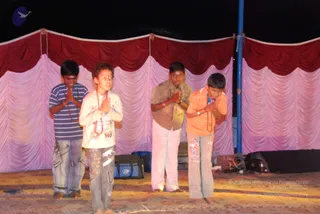 Four boys stand barefoot on a carpeted stage in front of a draped backdrop of red and pink curtains at night. They stand with palms pressed together in anjali namaskar. The boy on the left wears a blue and white striped top and jeans; the second wears a cream top with red beads around his neck and patterned trousers; the third wears a beige vest over a white shirt and white pants; the fourth on the right wears an orange top with a red mala and light trousers. Equipment bags and stage gear sit at the back edge of the stage.