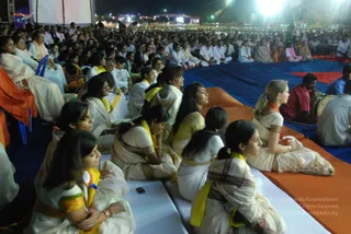 A large night gathering of seated devotees on blue and orange mats outdoors, viewed from the side. Women in white and cream cotton saris with yellow or gold borders and orange shawls sit in the foreground, several with hands folded or resting on their faces. Behind them rows of men and women in white kurtas and white clothing fill the audience area under bright stage lights. At the right edge a drummer in a red garment sits beside a drum. No a person candidate is clearly visible in this crowd view, and there is no visible figure on stage. The scene is a satsang-style audience at night with a stage and tents in the background.