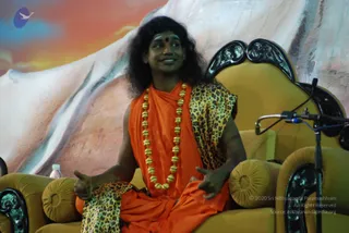 A man seated on an ornate gold-upholstered throne-style chair with black carved wooden arms, speaking on stage in front of a large backdrop of a snowy white mountain under a warm orange-blue sky. He wears an orange-saffron shawl draped over his shoulders and body, a leopard-print wrap over one shoulder and across his lap, and a long necklace of gold beads, and has long dark hair and a red bindi on his forehead. He gestures with both hands while smiling and looking upward, with a microphone on a stand beside him and a water bottle on the stage. Appearance-based a person with long swept hair, saffron clothing and gold beaded necklace, seated in a throne-style chair at a satsang-style talk.
