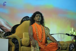 A seated man in an ornate golden-yellow throne chair with black carved wooden arms, on a stage before a colourful blurred backdrop of blue, green and yellow. He wears an orange saffron robe, a leopard-print shawl over one shoulder, a long gold bead necklace and a red bindi, with long swept-back dark hair. A microphone on a flexible stand sits beside him. Swamiji (probable): a sannyasi in orange with a leopard-print wrap, long hair and gold beads, seated on a throne during a satsang talk.