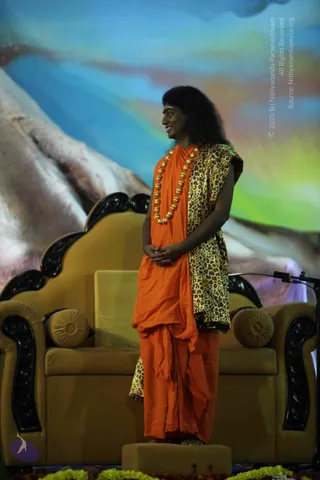 A standing man in orange saffron robes wrapped over a leopard-print shawl stands on a low golden platform on a stage. He wears a long necklace of large golden beads and has long, swept-back dark hair; his hands are clasped in front of him as he smiles and looks to the side. Behind him is a gold upholstered sofa-style throne with black carved armrests and round cushions, in front of a painted backdrop of blue and green sky over pale mountains. A microphone stand is visible at right, and marigold garlands line the stage edge at the bottom.