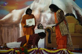 A stage with a large painted mountain backdrop in orange and blue tones. A woman in an orange sari with a leopard-print shawl stands at right, holding an open book, beside a gold-upholstered armchair with carved dark wood trim. A man in an orange robe stands at centre holding a white paper bag printed with a tree. A man in a maroon kurta and white dhoti lies prostrate on the floor in front of a white pedestal holding a brass bowl, in an act of obeisance. Marigold and flower garlands in yellow, orange and purple border the stage floor in front.