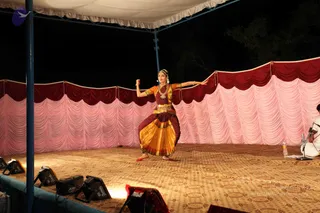 A female Bharatanatyam dancer performs on an outdoor stage at night under a white canopy, in front of a pink and maroon draped backdrop. She wears a maroon blouse with orange sleeves, a yellow and maroon pleated skirt, a gold waist belt, a gold necklace and a flower-decorated hair bun, and holds a small red object in one raised hand while extending the other arm. Stage lights sit at the front edge of the carpeted platform, and a seated musician in white at the right edge is partly visible beside a microphone and water bottle.