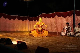 A female classical dancer performs on a stage at night under a tent, seated in a half-squatting pose with hands on her hips. She wears an orange and gold costume with a pleated fan-like skirt, a gold waist belt, layered white pearl necklaces and a headpiece. Behind her, two men in white traditional dress sit on the floor playing a violin and a percussion instrument at microphones. The backdrop is draped in pink and maroon fabric, the floor is covered with a patterned carpet, and stage lights glow at the bottom edge.