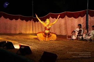 A female classical dancer performs at night on a carpeted stage in front of a draped pink and maroon backdrop. She sits in a cross-legged pose with both arms raised outward and palms open, wearing a gold and orange pleated costume, a gold necklace, and a head ornament. Two seated musicians to the right play a violin and accompany her; one wears a white kurta and the other a white shirt. Stage lights and speakers frame the lower foreground.