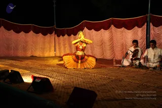 A night performance on an open stage with a patterned carpet, draped pink and maroon curtains behind, and stage lights in the foreground. A dancer in a gold and red costume with a jewelled headpiece and necklaces sits cross-legged in a namaskar pose with palms joined at chest level. Two musicians in white kurta and dhoti sit to the right beside microphones; one plays a violin and the other plays a percussion instrument. No a person candidate is visible.