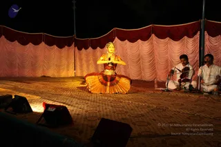 A female classical dancer in a golden and maroon costume with a gold headpiece and jewellery sits cross-legged on a patterned carpet on an outdoor stage at night, hands joined in namaste in front of her chest. Behind her, a draped pink and maroon curtain backdrop lines the stage. To the right, two male musicians in white kurta-style clothing play a violin and sit before a microphone stand. Stage lights and speakers are visible at the lower left. No a person candidate is visible in the frame.