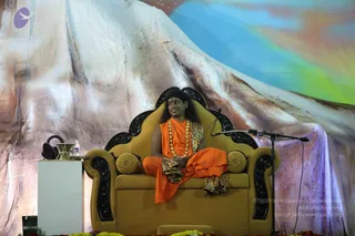 A man in orange saffron robes sits cross-legged on an ornate golden-yellow throne with black carved wooden arms and back. He wears a long rudraksha mala and a gold necklace, a leopard-print shawl draped over one shoulder, and a red bindi. Long dark hair falls past his shoulders. A microphone stand stands beside him, and a small white side table holds a water bottle and a bowl. Behind him is a large painted backdrop of snow-capped mountains against a blue and green sky, and marigold flowers line the stage base. Swamiji (probable) is seated on the throne, identified by orange robes, rudraksha mala, leopard shawl and long hair.