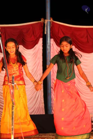 Two young girls stand hand in hand on a stage in front of draped red and pink curtains, with a microphone stand at left. The girl at left wears a red blouse with gold zari and a yellow-orange long skirt, with gold necklace and earrings. The girl at right wears a green blouse, a gold necklace with a red bead strand, and a peach-pink skirt with small gold motifs; both have gold bangles and red bindi marks. They appear to be performing a stage dance. No a person candidate is visible.