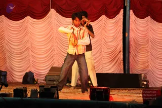 Two young boys perform on a stage draped with pink and red pleated curtains. In the foreground a boy in a yellow-and-orange plaid shirt over white sleeves and dark jeans stretches his arms out with his hands open, while a taller boy behind him in a dark shirt and white trousers makes a hand gesture near his face. Monitor speakers, a bag and a small cabinet sit at the stage edges, and a microphone stand is visible at the left.