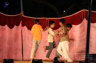 Four boys perform on a stage at night, dancing or moving energetically in front of a backdrop of draped red and pink fabric with scalloped valances. The boy on the left wears an orange and white striped polo shirt and light grey trousers. The boy in the centre wears a cream long-sleeved top with dark patterned shorts and is barefoot. The boy on the right wears a khaki short-sleeved shirt and white trousers, and another boy in a blue striped shirt is partly visible behind. Stage lights sit at the bottom of the frame and a tall blue pole stands at the right edge.