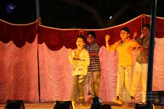 Four young boys perform on an outdoor night stage in front of draped red and pink curtains with a dark tree silhouette above. The boy at left in a cream long-sleeved top with a red bindi and a red-and-brown cord, arms bent, mouth open mid-chant or cheer. Next to him a boy in a navy and white striped polo and grey trousers stands behind. At right a boy in a bright yellow-orange striped shirt with a beaded necklace raises a fist, and at far right a boy in an olive-green shirt and white trousers lifts his arm. Stage lights glow at the bottom edge and a microphone stand stands at right.