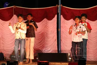 Four young boys perform on a stage at night in front of draped curtains, with red valances above and pink and rose-coloured pleated drapes below. The boy on the far left wears a yellow and orange striped shirt, white undershirt and blue jeans. The second boy wears a dark patterned shirt and cream trousers with a small embroidered design. The third boy wears a white and pink checked shirt with a lanyard and jeans, and the boy on the far right wears a red and white t-shirt with blue jeans. All four are barefoot or mid-motion, with hands raised in a dance-like gesture, and a small red tilak mark is visible on a couple of foreheads. A stage monitor speaker and cables sit at the front of the platform, and a blue pole stands behind the performers.