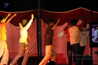 Photo of a stage performance at night in front of draped red and maroon curtains. Six young male performers in casual clothes, including an orange striped shirt, a white shirt with a patterned collar, a dark brown printed shirt, a red and white raglan shirt and a plaid shirt, strike energetic poses with arms raised and bodies turned. A small monitor on the right shows the stage. No a person candidate is visible; the performers are dancers.