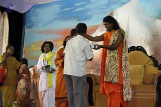 A stage event with a painted backdrop of a blue sky, orange clouds and a white mountain. On the right, a long-haired man in orange robes and a leopard-print shawl smiles while reaching toward a seated man in a white checked shirt and grey trousers who holds a small object in his hand; the robed figure appears to be presenting or handing something over. A man in white kurta and a yellow shawl stands behind, holding a small black device. A woman in a yellow sari stands at the left edge, and another person in orange robes stands behind the exchange. Gold-upholstered chairs with carved black frames sit on the right. Swamiji (probable): the long-haired figure in orange with a leopard-print shawl, presenting an object on stage.