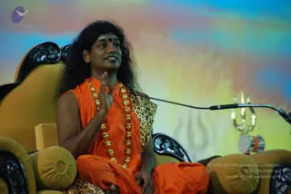 A man with long, swept-back dark hair sits cross-legged on an ornate gold-and-black throne-style chair, wearing an orange sleeveless robe with a leopard-print shawl draped over one shoulder and a long gold bead necklace. He raises one hand mid-gesture while speaking, and a microphone stand is in front of him. Beside him stands a gold trident and a small drum on the right. Behind him is a colourful blue, yellow and pink backdrop with a faint Om-like symbol. The appearance-based label: a person, seated on a throne during a discourse.