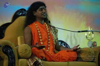 A man with long, swept-back dark hair and a red bindi sits cross-legged on an ornate golden-yellow throne with carved black armrests and round cushions. He wears an orange saffron robe draped over his body, a leopard-print shawl, and a long necklace of gold beads. His hands are open, palms up, as he speaks. A microphone on a stand sits beside him, and a silver trishula with a small damaru drum rests on the right armrest area. The background is a soft, multicoloured wash of blue, orange and yellow, with a faint Om symbol. Swamiji (probable): long swept-back hair, saffron robe and ornate gold necklace on a throne, consistent with the page's Swamiji claim.