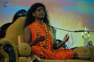 A seated man speaks during a satsang on a gold-upholstered throne with carved black wooden armrests. He wears a saffron-orange shawl and dhoti, a leopard-print cloth draped over the throne, long gold bead garlands, and a dark bindi. His long, swept-back dark hair falls past his shoulders, and he gestures with both hands while looking up. A microphone stand is in front of him, and a silver-and-gold trishula stands on a small drum-shaped stand at right beside a painted sunset-coloured backdrop. Swamiji (probable): his long swept-back hair, saffron clothing and gold bead garlands match the presentation cues, and he sits on a throne as the speaker. Only one person is visible.