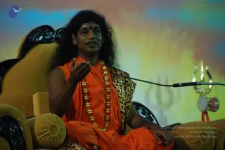 A man with long, voluminous dark hair and a red bindi sits on an ornate golden throne-style chair, speaking and gesturing with his right hand. He wears an orange saffron robe and a long necklace of large golden beads, with a leopard-print cloth draped over the chair back. A microphone stand is in front of him, and to the right a golden trident with a small damaru drum hangs against a multicoloured backdrop with a faint trident motif.