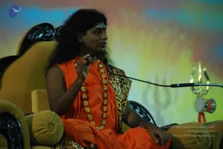 A seated man in saffron-orange robes speaks into a microphone while gesturing with his right hand during a talk. He wears a long heavy necklace of gold beads, has long swept-back dark hair and a red mark on his forehead, and sits in an ornate yellow upholstered throne chair. A leopard-print cloth is draped over his shoulder. Behind him a colourful painted backdrop shows a faint trishul design, and a silver trishul with a small drum hangs at the right edge. The compact photo shows a single person indoors.