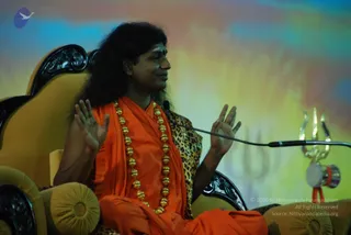 A photograph of a long-haired man seated on an ornate dark-wood throne with gold-cushioned armrests, speaking during a satsang. He wears an orange saffron shawl over his body and a long gold bead necklace, and his palms are raised in gesture. A microphone on a stand is at his side, and a golden trident with a small damaru hangs from the mic stand on the right. The backdrop is a soft blue, orange and yellow gradient with a faint trident motif. Swamiji (probable): the long swept-back hair, saffron clothing, gold necklace and trident setting match the page's talk, but the face is not used for identification.